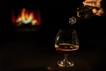 Woman pours cognac into a glass on the background of the fireplace.