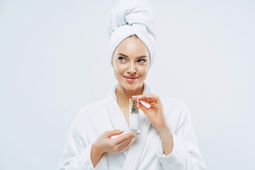 Thoughtful beautiful woman with clean fresh skin, recommends cosmetic product, feels relaxed after taking bath, wears dressing gown, wrapped towel on head. Beauty, wellness, body care concept