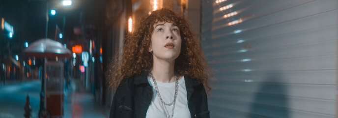 Young woman with curly hair in white t-shirt and black jacket is walking on the street at night,...