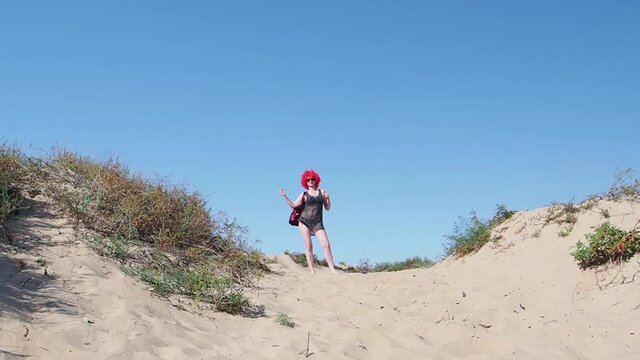 A posh crazy mid-aged woman in a shiny swimsuit, mesh tunic and red wig walked to the sandy beach. Woman dancing and celebrating hallowin on the seaside. Slow motion