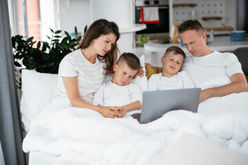 Obraz premium Beautiful parents with kids enjoying at home. Young family watching movie on lap top.