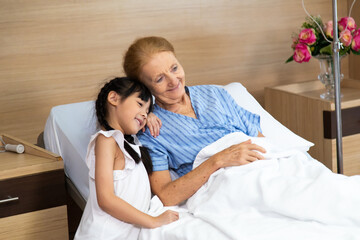 Fototapeta premium Happy elderly patient lying on hospital bed with grandchild visitor. grandmother and granddaughter enjoying talking together.
