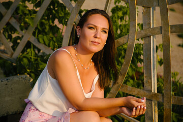 Portrait of a girl sitting on the background of metal railing on a clear sunny day and looking somewhere to the side.