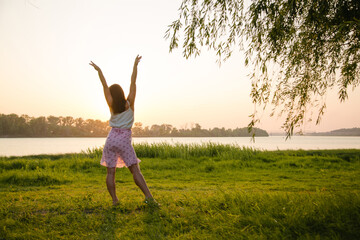 A beautiful girl raised her hands on the bank of the river, in the rays of the setting sun. Fresh air, unity with nature.