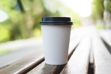Obraz na płótnie Canvas A paper cup of coffee to go on bench with blurred nature and sun background. Take-away cup with plastic lid.