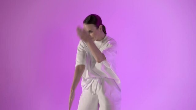 Girl dancing hip hop in white sport clothes moves hands on violet background. Studio shot of young happy girl professional dancer in white clothes and black boots