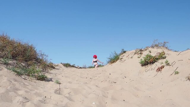 woman wearing red wig, shiny swimsuit coming out of the hill and walking towards the camera while walking at the beach. crazy middle aged woman celebrating hallowin - video in slow motion