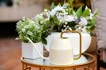 Home decor on the terrace of blooming flowers in white pots.A white decorative watering can and home plants stand on a glass table.Selective focus.