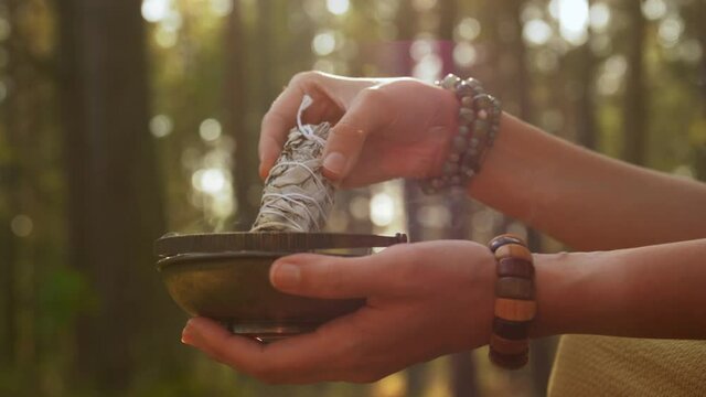 occult science and supernatural concept - young woman or witch with smoking white sage performing magic ritual in forest