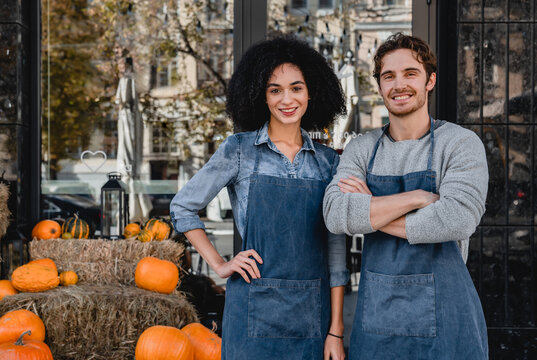Portrait of young man and woman cafe owners standing outdoor front of their cafe with decorating pumpkins preparing for halloween. - Powered by Adobe