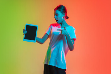 Showing tablet's screen. Young caucasian girl's portrait on gradient green-orange studio background in neon light. Concept of youth, human emotions, facial expression, sales, ad. Beautiful teen model.
