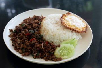 Side view of Rice topped with stir-fried beef and basil with fried egg in the white plate. Background is tile black color.