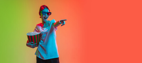 Wearing 3D-headset, pointing with popcorn. Young caucasian girl's portrait on gradient green-orange studio background in neon light. Concept of youth, human emotions, facial expression, sales, ad