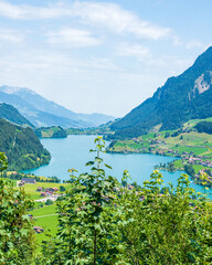 Panorama d'un lac bleu turquoise avec un petit village encastré entre des montagnes, avec de la végétation au premier plan, un jour ensoleillé avec des nuages