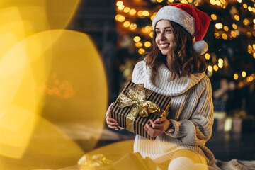 Pretty woman in warm sweater, socks and christmas hat, sitting on the floor at home with gift boxes