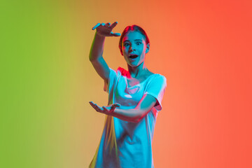 Shocked holding blank space. Young caucasian girl's portrait on gradient green-orange studio background in neon light. Concept of youth, human emotions, facial expression, sales, ad. Beautiful teen