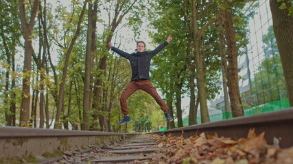 Naklejka premium Playful happy man teenager, student, rejoices at success. A young guy funny bouncing in the park and jumps with happiness against the backdrop of a beautiful sunset. Slow motion. on the front