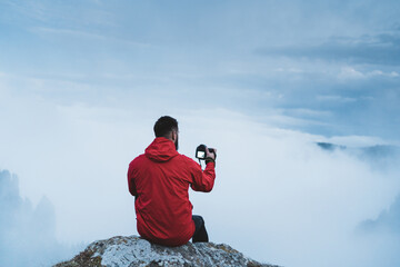 Young bearded hiker wearing red jacket taking photos at mountain with fog and mist surrounding...