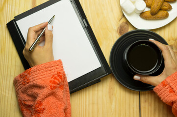 Female hands in a white sweater on a wooden table hold a pen in front of a notebook. Wooden table with a cup of coffee, a light snack and a notepad for writing ideas.