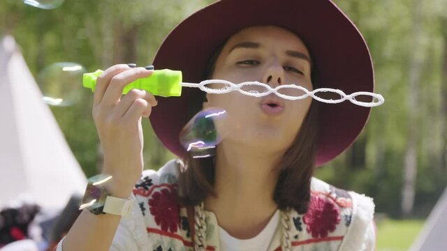 Front-view closeup of joyful young woman in hat blowing soap bubbles outdoors laughing out loud in daylight with summer forest in background