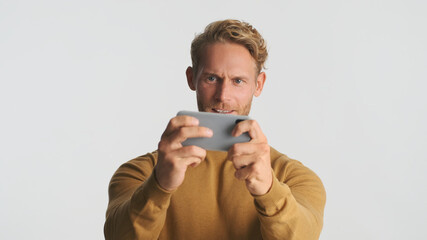 Attractive emotional bearded man intently playing in game on smartphone over white background. Gamer concept