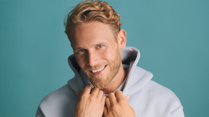 Handsome smiling bearded man in hoodie happily looking in camera and smiling isolated on blue background. Male model