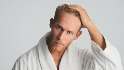 Attractive bearded man in bathrobe looking serious preening on camera over white background