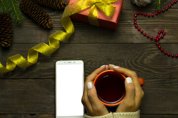 Female hands in a white sweater on a wooden table hold a hot mug with a drink. On the table is a smartphone with a white screen. Wooden table with festive decoration, gift and decoration.