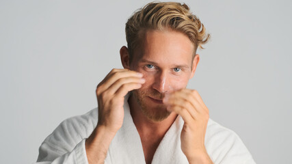 Attractive bearded man in bathrobe applying cream over white background. Male model in studio