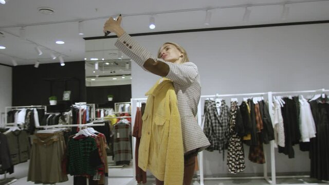 Young attractive woman is taking a selfie with yellow jacket at clothing store, tracking shot, 360 degree