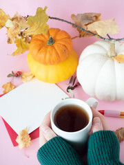 Hands holding cup of tea near white paper on red envelope with autumn leaves and pumpkins around