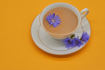 A Cup of drink stands on a saucer on a yellow background, several chicory flowers are lying next to it.