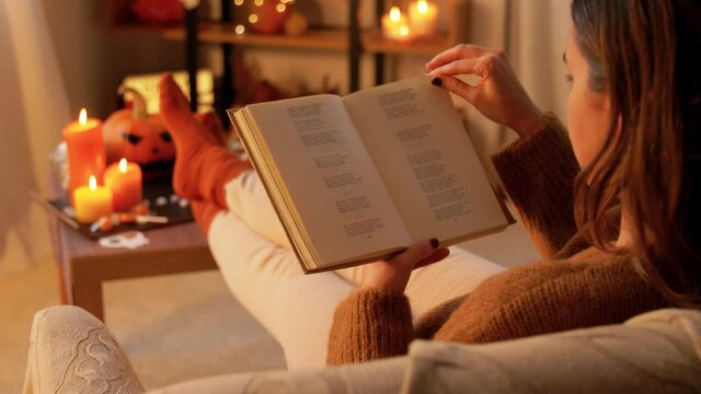 halloween, holidays and leisure concept - young woman reading book and resting her feet on table at cozy home
