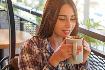 Portrait of young beautiful woman enjoying cup of cocoa w/ marshmallow topping at restaurant. Joyful female experiencing satisfaction from drinking hot beverage on cold autumn day. Close up copy space