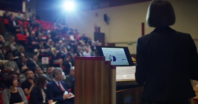 Back view of female professor is holding a speech to the audience during business economics and finance congress event in an auditorium. 