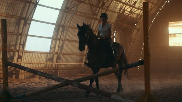 Female jockey on the horse is running in the hangar and jumps over obstacle.