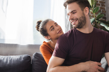 happy woman embracing happy boyfriend and looking at him while sitting on sofa at home