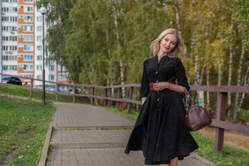 Smiling stands and leans on the railing, an adult with a kind look in black clothes, in the fall against the background of trees, a blue sky.