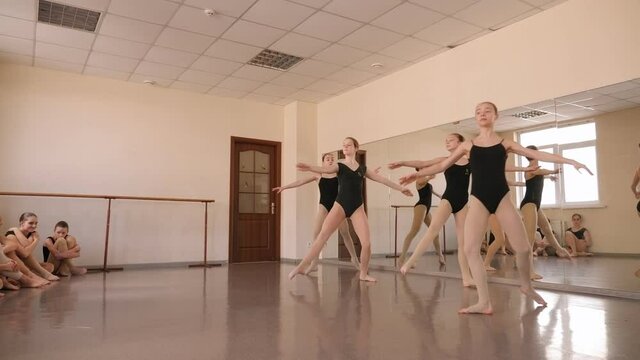 Four flexible young ballerinas are jumping in the splits in a ballet class, with ballerinas sitting in the background.