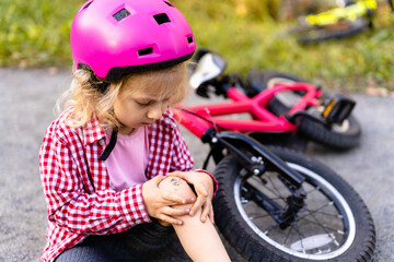 Fototapeta na wymiar little blonde girl fell off her bike and holds her bruised leg with her hands, sad expression on her face