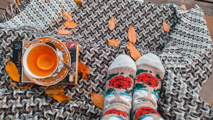Feet of a girl in socks in autumn sitting on the veranda with a book, tea, autumn leaves