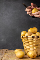 Obraz na płótnie Canvas Close-up of basket with yellow potatoes and woman's hands with paring knife, on rustic table, with black background, in vertical, with copy space