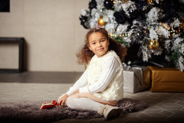 Happy smiling mixed girl with dark hair sitting on the carpet. Christmas tree in the background.