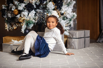 Happy smiling mixed girl with dark hair sitting on the carpet. Christmas tree in the background.