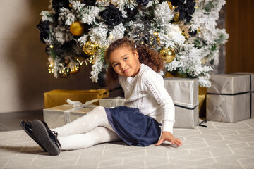 Happy smiling mixed girl with dark hair sitting on the carpet. Christmas tree in the background.