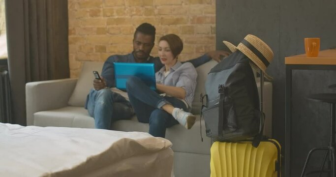Packed luggage standing in bedroom or hotel room with blurred interracial couple surfing Internet on laptop at the background. Happy young man and woman checking flight time online.