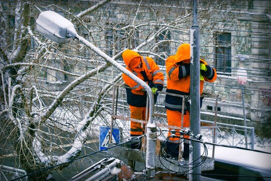 Two electricians in orange overalls on a lift work on top of an electric pole with cable lines against the backdrop of city buildings in winter.