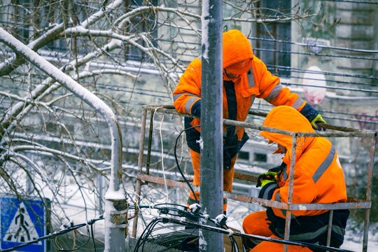 Two electricians in orange overalls on a lift work on top of an electric pole with cable lines against the backdrop of city buildings in winter.
