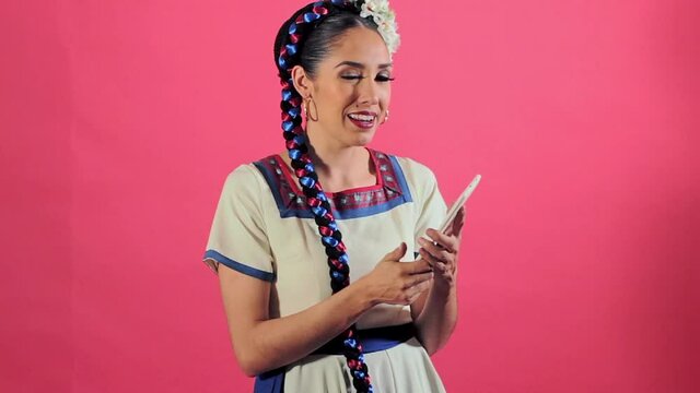 Joven mujer mexicana latina en traje regional folcl&oacute;rico en fondo rosa utilizando tableta para comunicarse redes sociales
