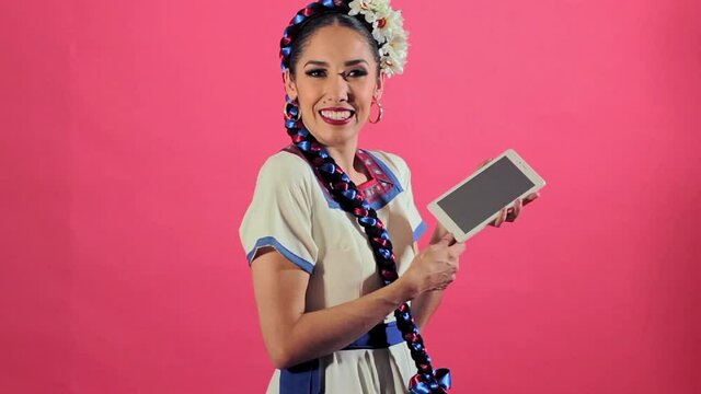 Joven mujer mexicana latina en traje regional folcl&oacute;rico en fondo rosa utilizando tableta para comunicarse redes sociales